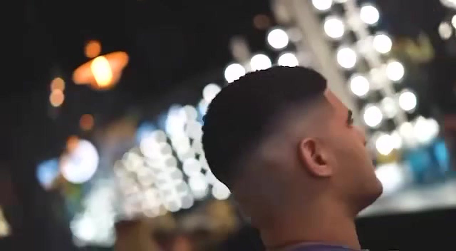 A client mid-fade seen from behind and to the side, lit by a wall of warm mirror bulbs against a dark room.